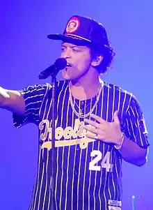 Bruno Mars down on one knee at the front of a stage, singing into a microphone, with someone in the audience holding his forearm. He is wearing black pants, a plaid button-up shirt, and a gray fedora.