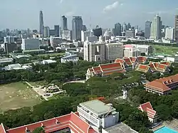 The campus of Chulalongkorn University, with many red-roofed buildings and trees; many tall buildings in the background