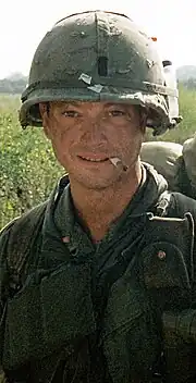 A man is at the center of the image looking at the camera. He is dressed in Vietnam-era military attire