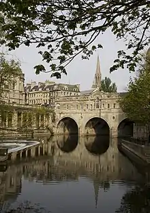 Yellow/Gray stone bridge with three arches over water which reflects the bridge and the church spire behind. A weir is on the left with other yellow stone buildings behind.