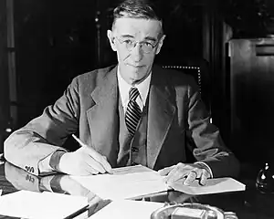 A man in a suit poses sitting at a desk, pen in hand. Papers and an ash tray are on the desk.